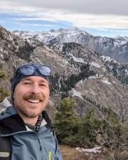 selfie of white man with light beard on a mountain with other mountains and blu esky
